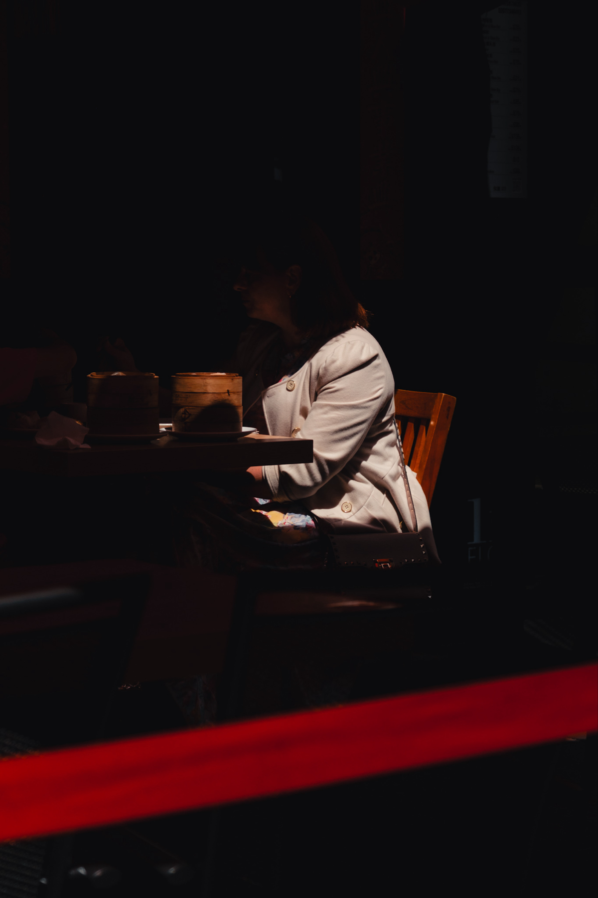 A person sitting at a table in a dimly lit setting, partially illuminated. The table has two wooden containers on it. A red stripe crosses the lower part of the image.