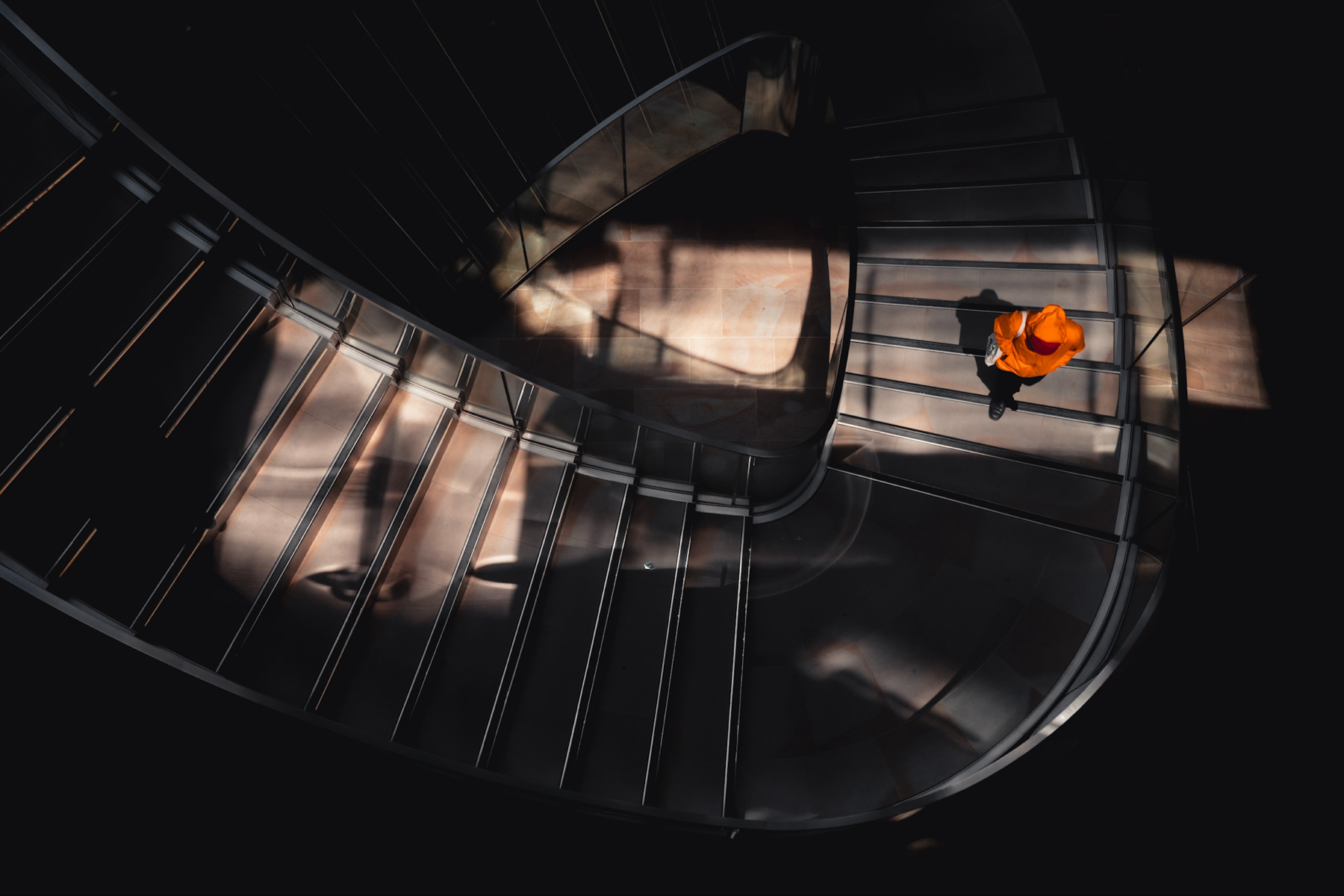 A person in a bright orange coat going up a dark spiral staircase shot from above 